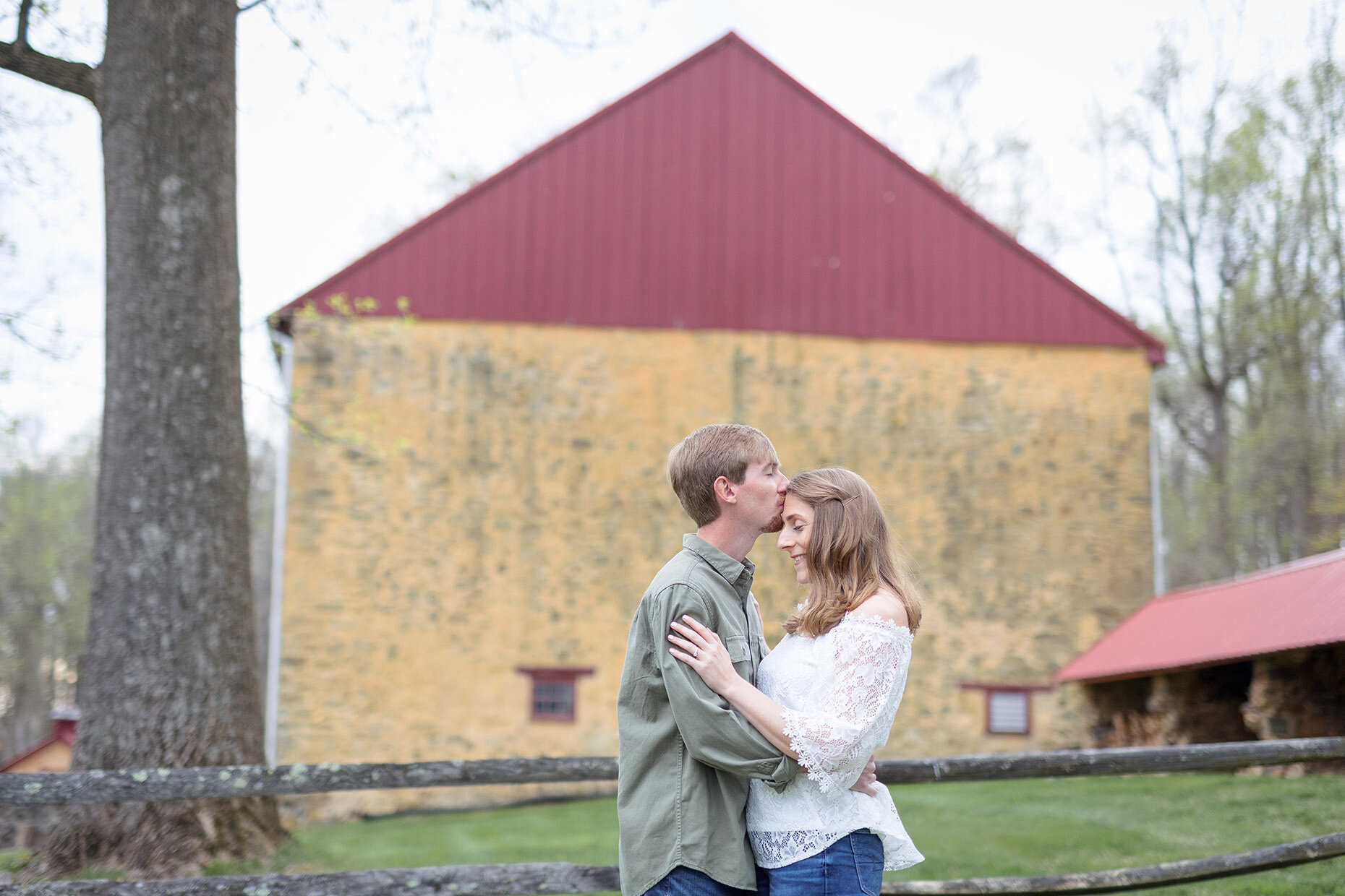 Couple kissing on forehead in front of the yellow barn at Hibernia Park
